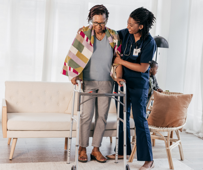 A caregiver assists an individual using a walker, with a colorful blanket draped over their shoulders.