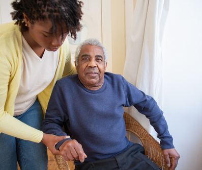 A caregiver in a yellow cardigan assists an elder sitting in a wicker chair.