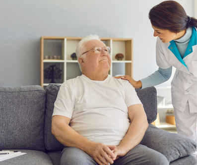 A caregiver in a white uniform places a comforting hand on the shoulder of an elderly man sitting on a sofa.