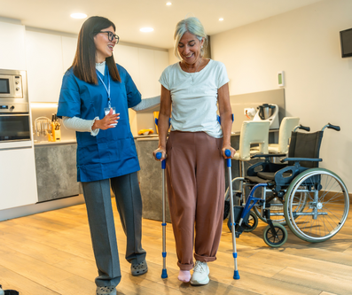 A caregiver in a blue uniform assists an individual using forearm crutches to walk. A wheelchair is visible in the background.