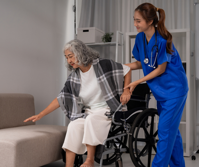 A caregiver in blue scrubs assists an individual in a wheelchair to reach for a sofa.