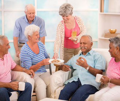 A group of seniors are gathered in a living room, sharing cookies and drinks. One woman is offering a plate of cookies to another, while a man holds a cookie and a woman holds a drink.
