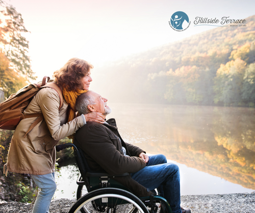 A person in a wheelchair by a calm lake, with another person standing behind and leaning in as if helping; autumn trees and misty sunlight fill the background.